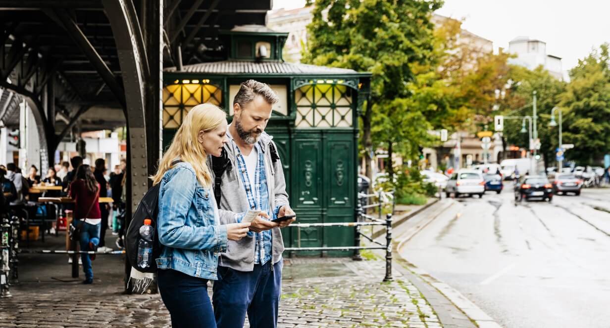 ein Mann und eine Frau stehen unter einer S-Bahnbrücke und schauen auf ein Handy.